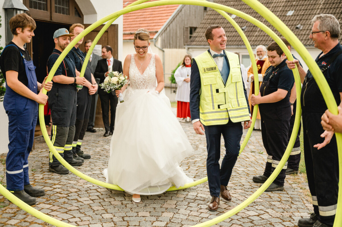 In einer feierlichen Atmosphäre im Freien gehen Braut und Bräutigam durch einen Kreis von Feuerwehrmännern mit Schläuchen.