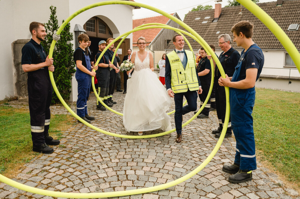 Braut und Bräutigam gehen durch große gelbe Schlauchbögen, die von Feuerwehrleuten auf einem Kopfsteinpflasterweg gehalten werden.