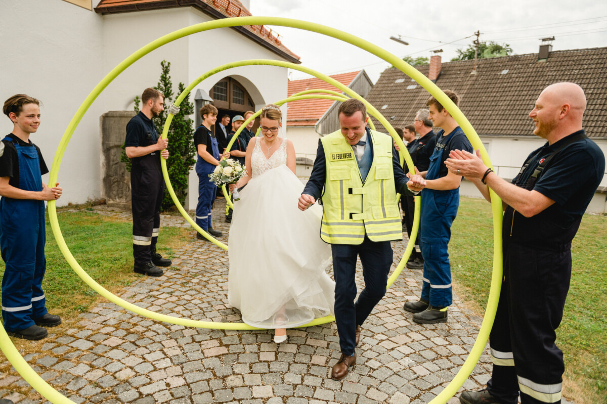 Braut und Bräutigam laufen im Freien durch Reifen, die von uniformierten Personen gehalten werden. Die Szene wirkt feierlich, mit klatschenden Zuschauern.