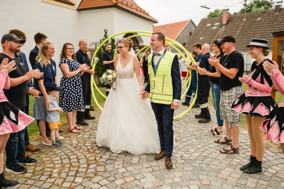Braut und Bräutigam gehen durch einen von Menschen gehaltenen Bogen aus Gartenschläuchen, während eine Gruppe klatscht und jubelt. Der Bräutigam trägt eine gelbe Sicherheitsweste.
