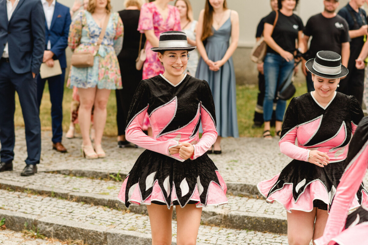 Zwei Darsteller in passenden schwarz-rosa Kostümen lächeln und halten Hüte. Im Hintergrund ist eine Menschenmenge in farbenfrohen Outfits zu sehen.