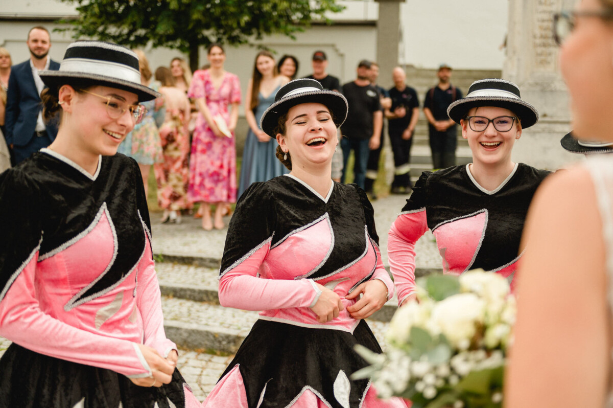 Drei Frauen in passenden schwarz-rosa Outfits und Hüten lächeln und lachen, im Hintergrund ist eine Gruppe von Menschen im Freien zu sehen.