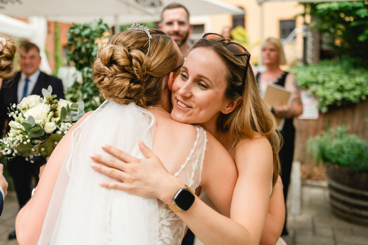 Eine Braut in einem weißen Kleid und Schleier umarmt eine Frau mit Sonnenbrille und Smartwatch bei einer Veranstaltung im Freien. Im Hintergrund sind weitere Personen zu sehen.