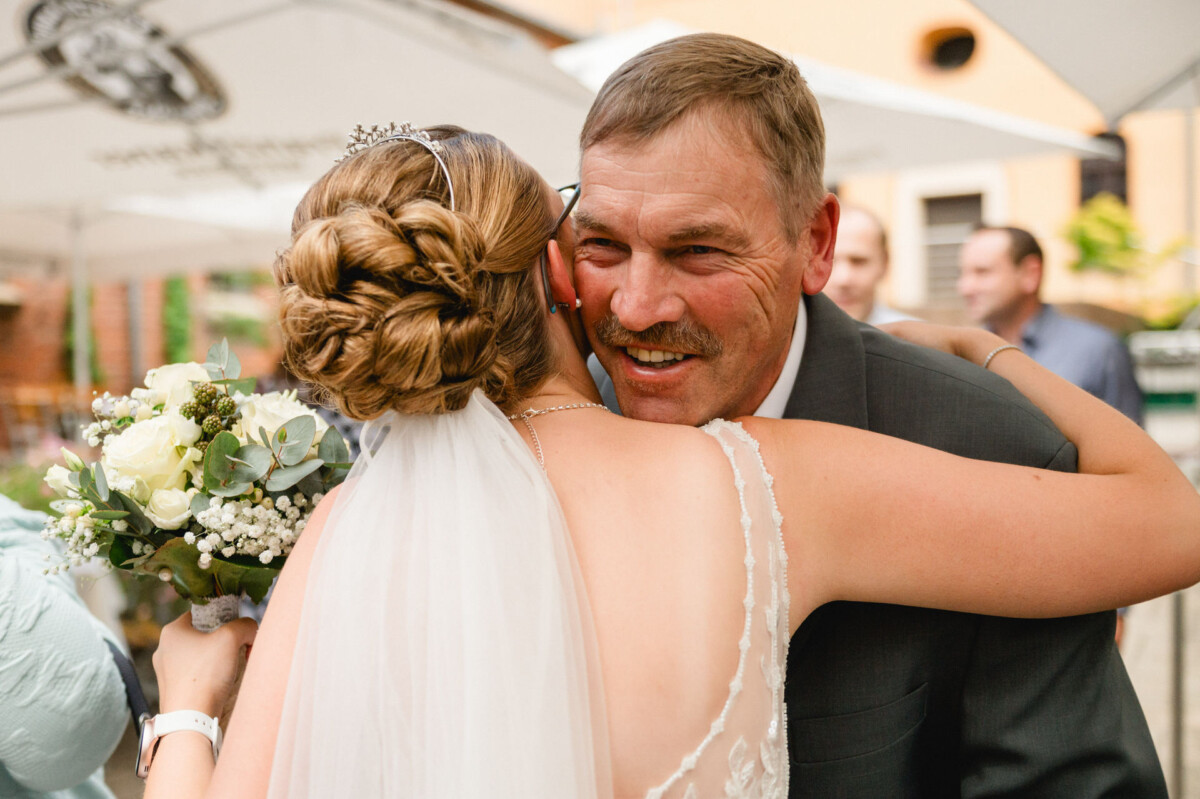 Eine Braut mit einem Blumenstrauß umarmt einen Mann im Anzug im Freien.