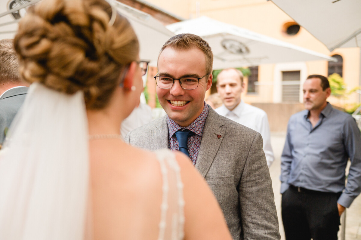 Ein Mann mit Brille und grauem Anzug lächelt eine Frau im Hochzeitskleid an, im Freien, im Hintergrund sind Menschen zu sehen.