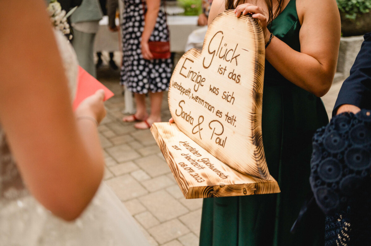 Person, die eine Holztafel mit deutschem Text hält und neben jemandem im Brautkleid steht. Bei der Veranstaltung scheint es sich um eine Hochzeit zu handeln.