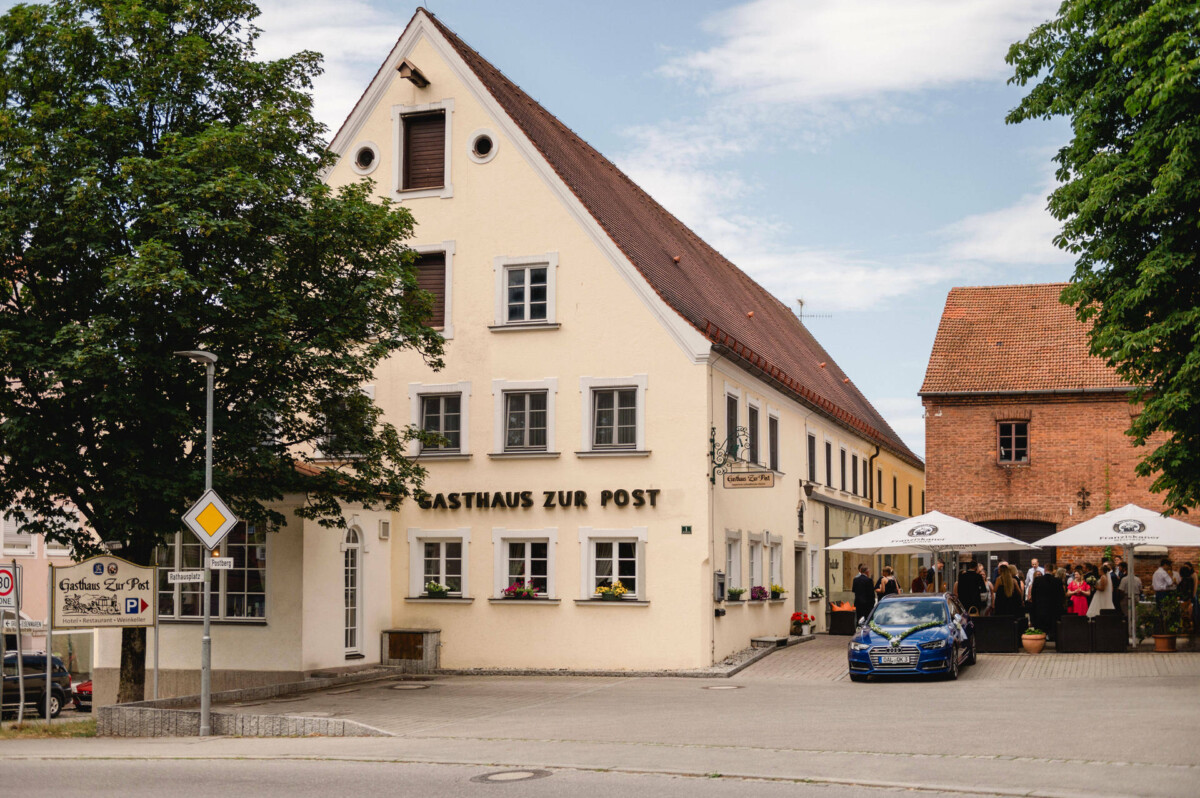Das Bild zeigt ein traditionelles deutsches Gasthaus mit dreieckigem Dach, das „Gasthaus zur Post“, vor dem sich Menschen versammelt haben. Davor ist ein geparktes blaues Auto zu sehen.