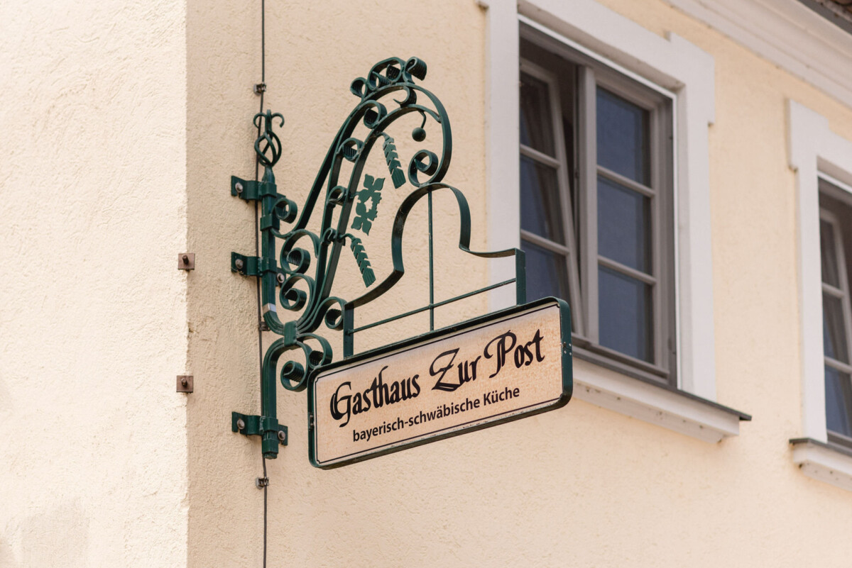 Schild „Gasthaus Zur Post“ mit bayerisch-schwäbischer Küche, an einem beigen Gebäude in der Nähe von zwei Fenstern angebracht.