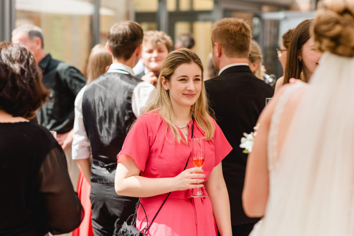 Eine Frau in einem rosa Kleid hält ein Glas, während sie bei einem geselligen Beisammensein inmitten einer Gruppe von Menschen steht.