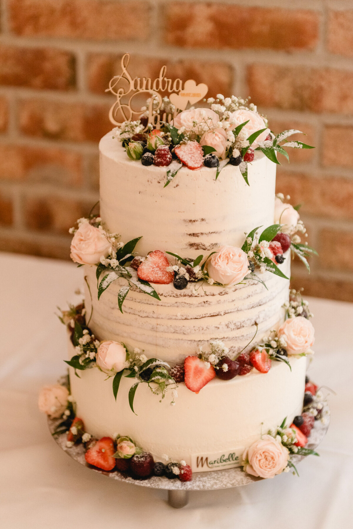 Dreistöckige weiße Hochzeitstorte mit rosa Rosen, Erdbeeren und grünem Laub auf einem Tisch. Zur Dekoration gehört ein Topper mit dem Text „Sandra & Gio“ und eine Backsteinmauer im Hintergrund.