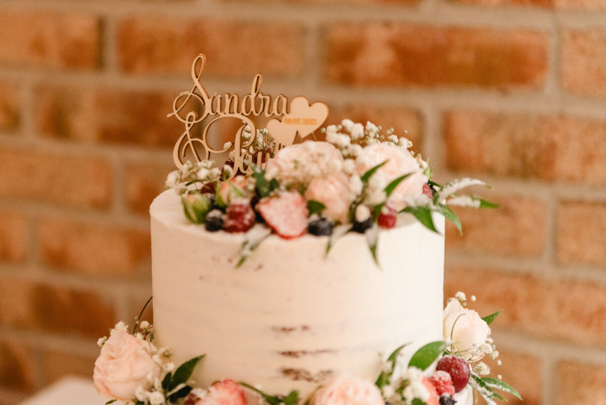 Eine zweistöckige Torte mit weißem Zuckerguss, Blumendekorationen und einem Holzaufsatz auf der oberen Etage vor dem Hintergrund einer Ziegelmauer.