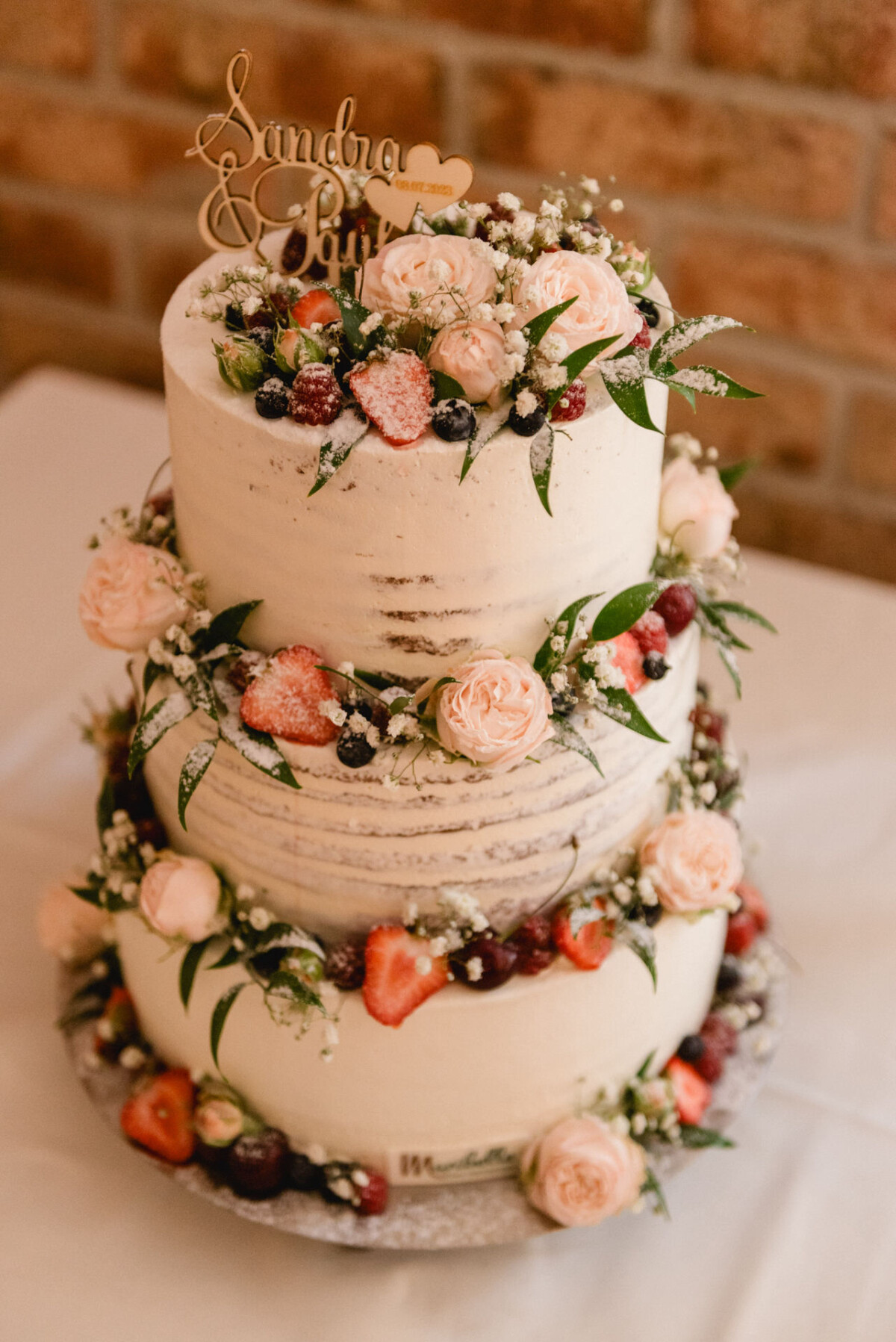 Dreistöckige Hochzeitstorte mit rosa Rosen, Erdbeeren und Grünpflanzen, gekrönt mit einem hölzernen „Sandra & Paul“-Schild.