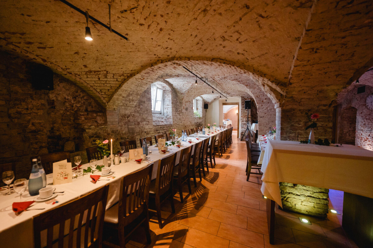 Ein langer Esstisch, gedeckt für eine Veranstaltung in einem rustikalen, gewölbten Backsteinraum mit Holzstühlen und stimmungsvoller Beleuchtung.