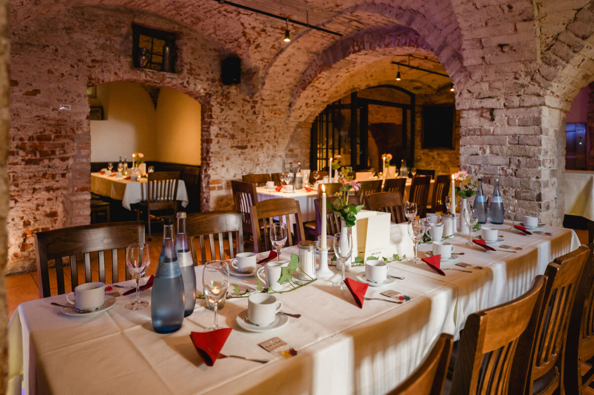 Ein schwach beleuchtetes Restaurant mit Backsteinbögen und Tischen mit weißen Tischdecken, Geschirr, Weingläsern, Wasserflaschen und roten Servietten.