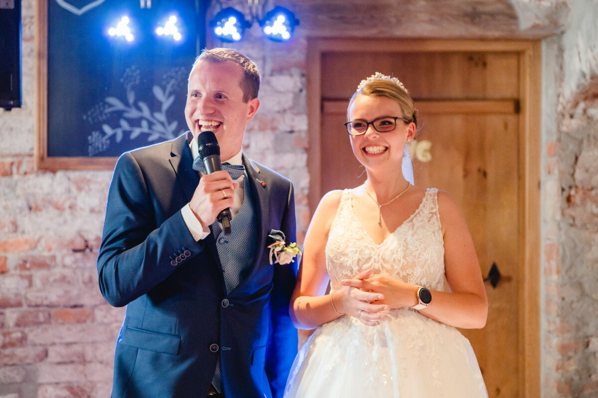 Ein Paar im Hochzeitskleid lächelt, während der Mann ein Mikrofon hält. Sie stehen in einem Raum mit Ziegelwänden, im Hintergrund blaues Licht und eine Holztür.