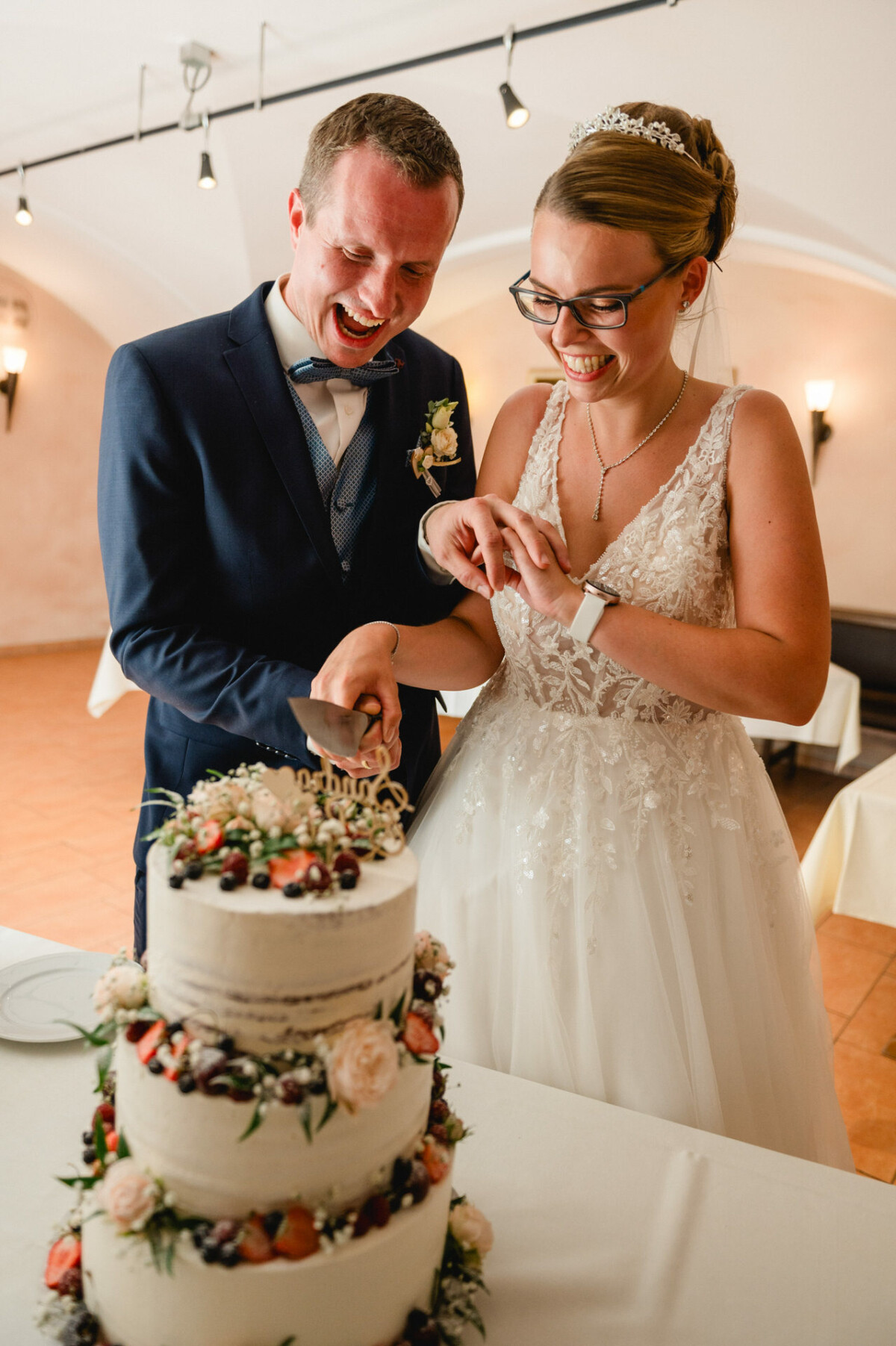 Braut und Bräutigam lächeln, während sie in einem Empfangssaal eine dreistöckige, mit Blumen und Beeren geschmückte Hochzeitstorte anschneiden.