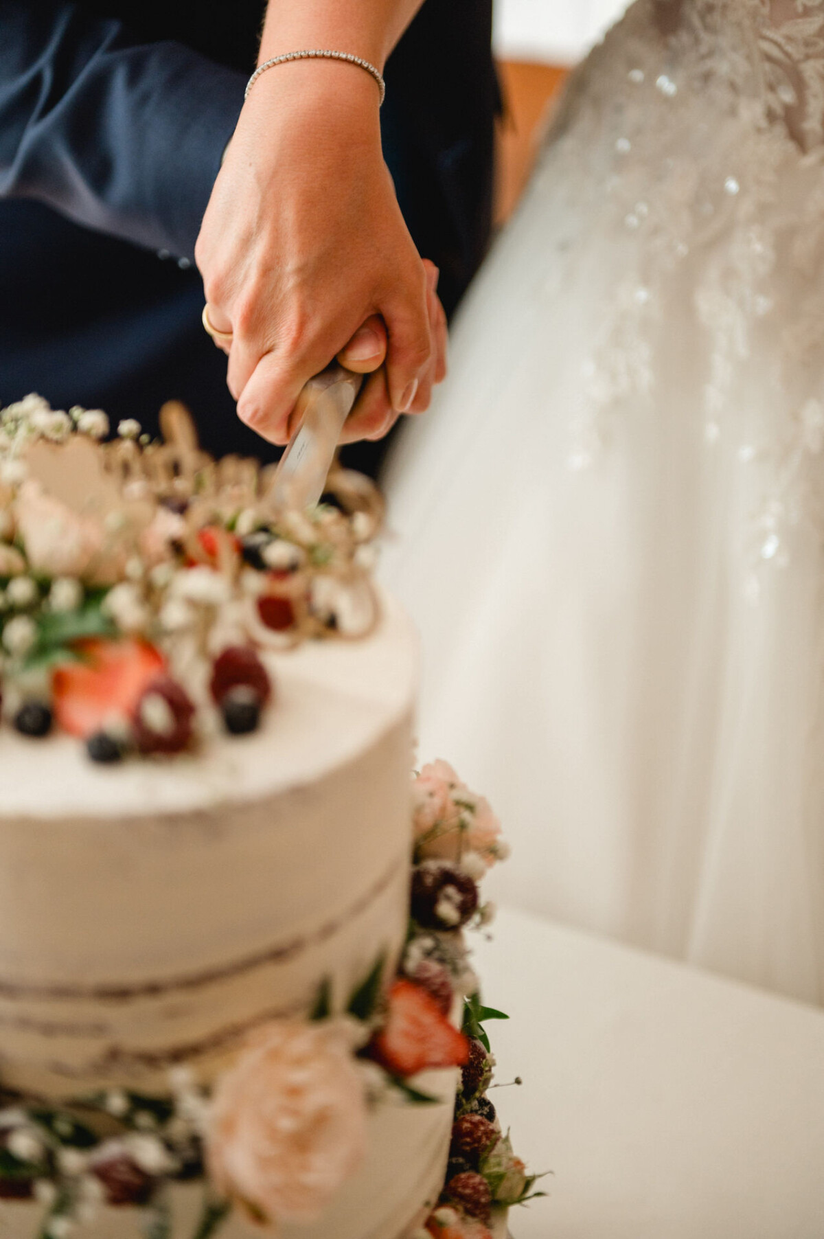 Ein Paar schneidet eine zweistöckige Hochzeitstorte an, die mit Blumen und Beeren geschmückt ist.