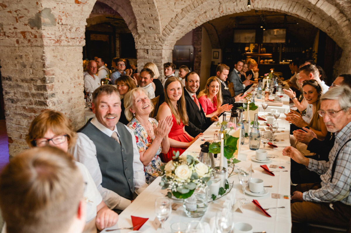 Menschen sitzen an einem langen Tisch in einem rustikalen Raum mit Backsteinwänden und lächeln und klatschen während einer Versammlung oder Feier.