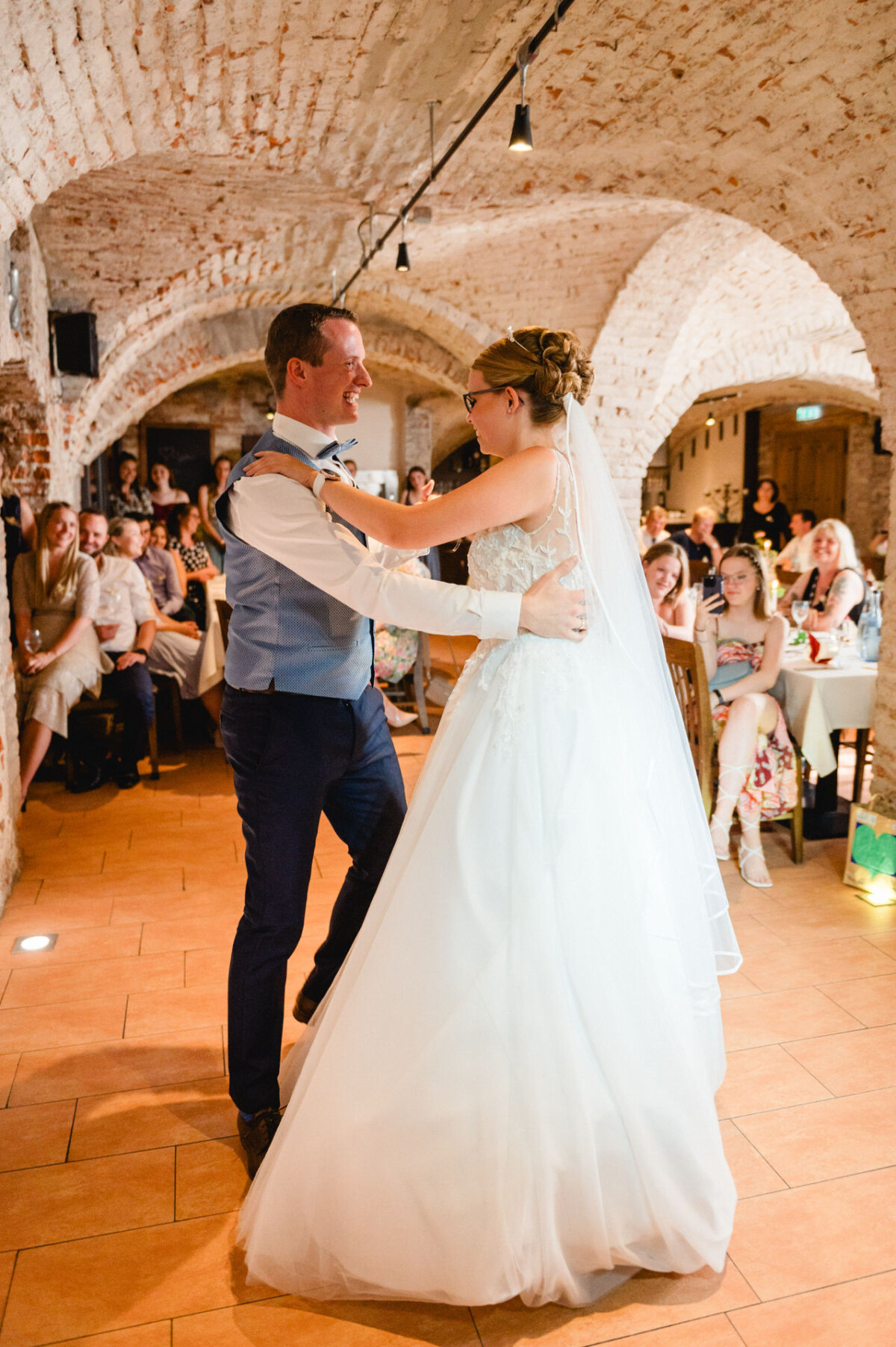 Braut und Bräutigam tanzen ihren ersten Tanz in einem Empfangssaal mit Backsteinwänden, umgeben von sitzenden Gästen.
