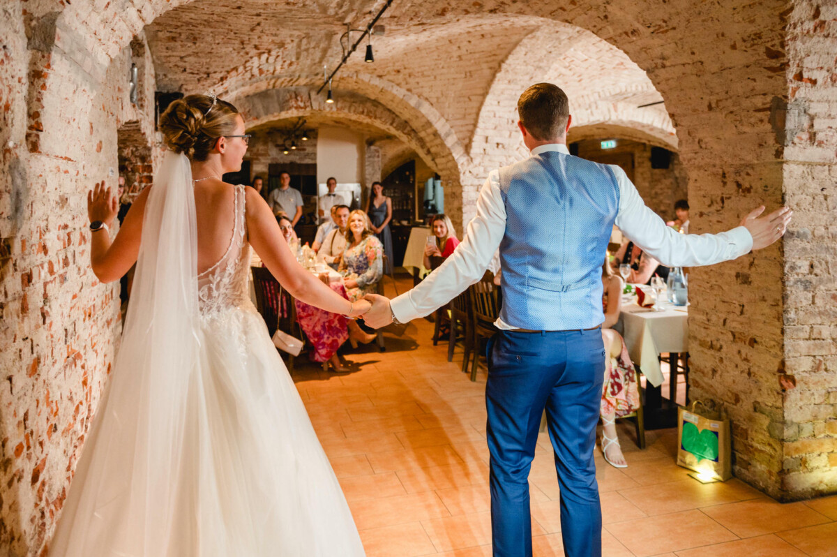 Braut und Bräutigam betreten Händchen haltend einen rustikalen, gewölbten Backsteinsaal und blicken auf die an den Tischen sitzenden Gäste.
