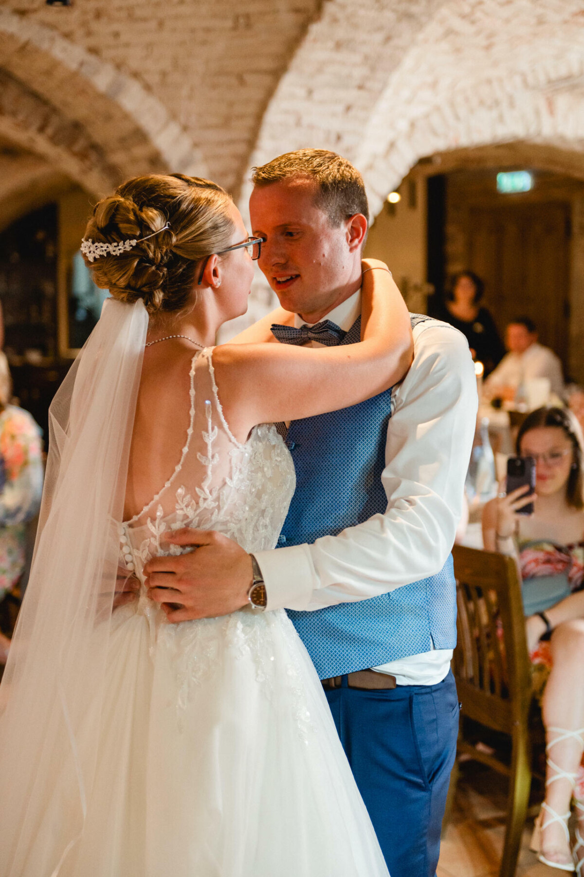 Braut und Bräutigam tanzen in einem warm beleuchteten Saal. Die Braut trägt ein weißes Kleid mit Schleier und der Bräutigam eine blaue Weste und Krawatte. Im Hintergrund sitzen Gäste, einige machen Fotos.