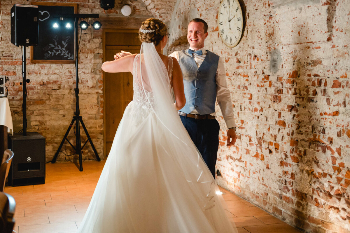 Braut und Bräutigam tanzen in einem rustikalen Lokal mit Backsteinmauern und einer Uhr. Die Braut trägt ein langes Kleid, der Bräutigam eine Weste und eine Krawatte.