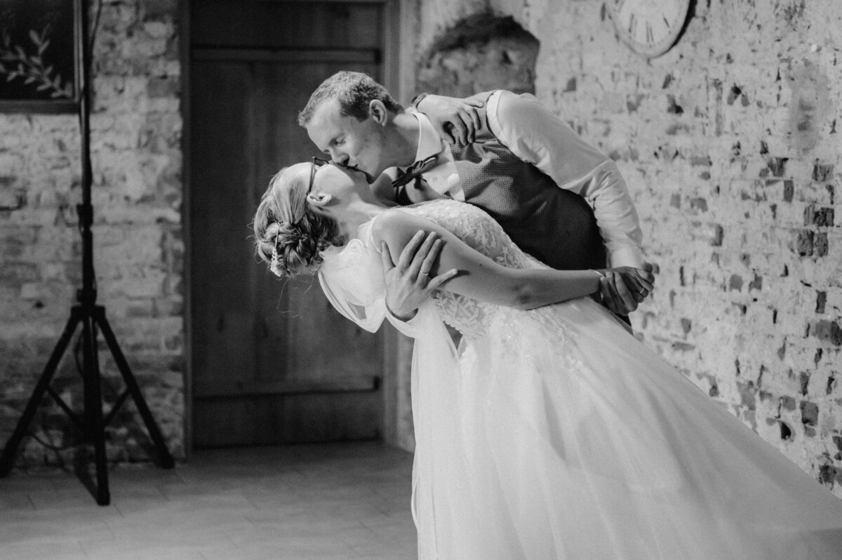 Ein Paar im Hochzeitskleid gibt sich einen romantischen Kuss, während es in einer rustikalen Umgebung tanzt.