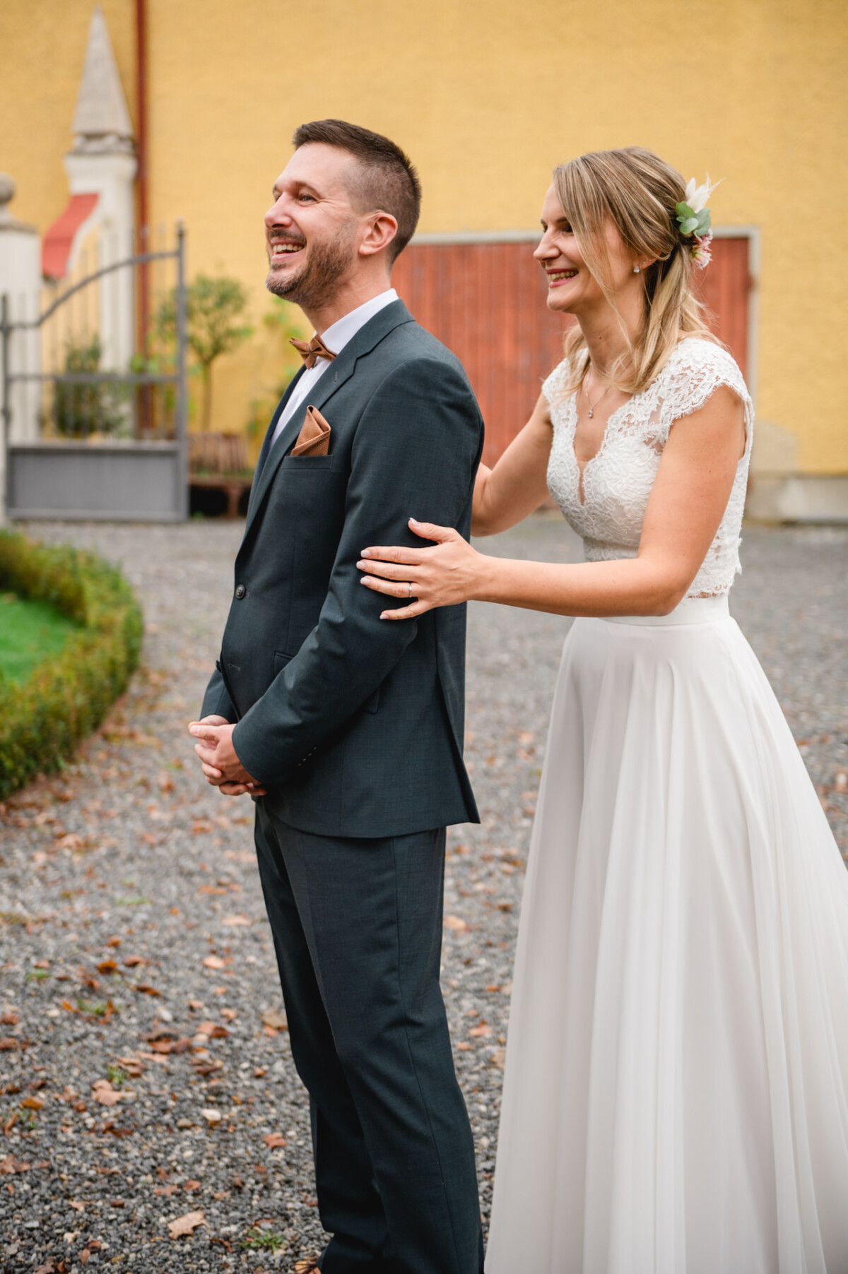 Eine Braut in einem weißen Kleid steht lächelnd hinter einem Bräutigam im dunklen Anzug und hat im Freien ihre Hände auf seinen Schultern.