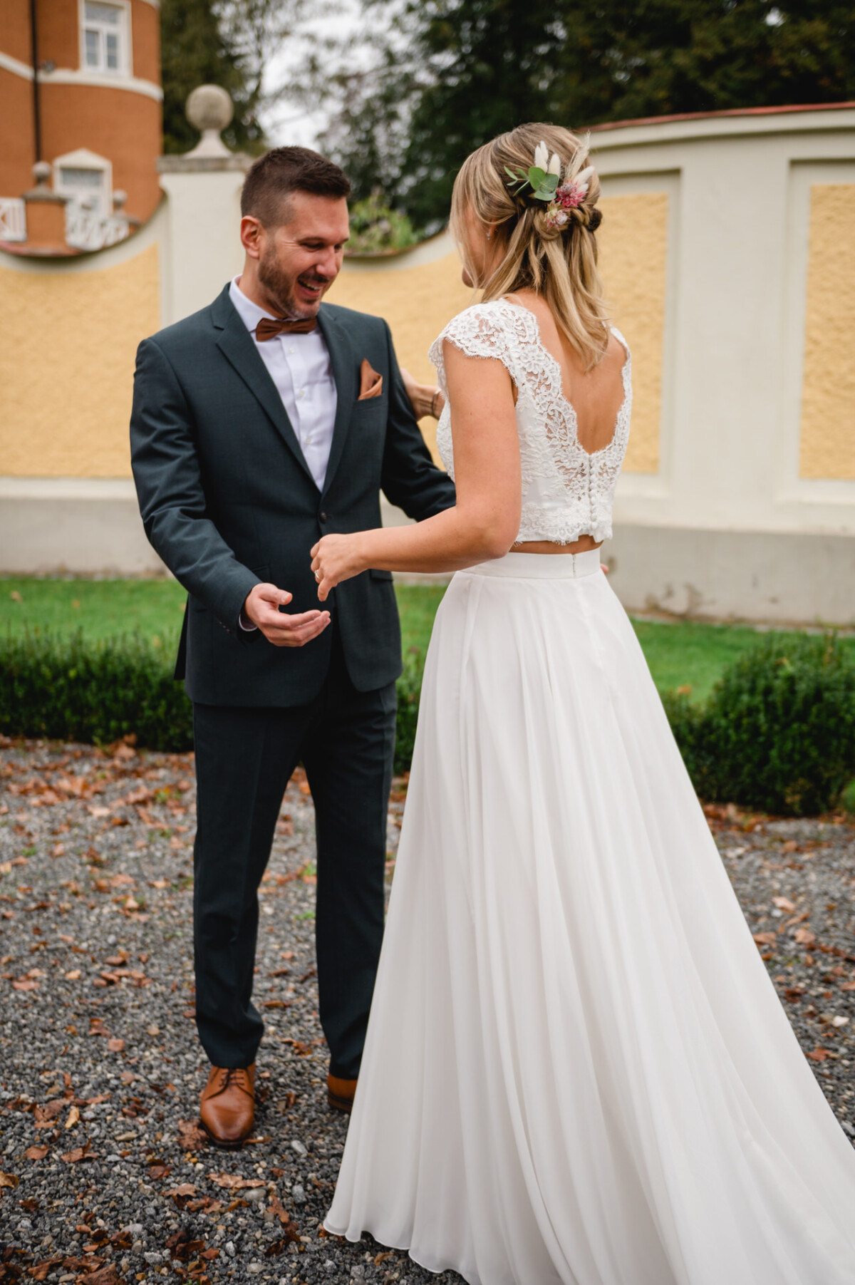 Braut und Bräutigam stehen sich im Freien auf einer Kiesfläche gegenüber. Der Bräutigam trägt einen dunklen Anzug und die Braut ein weißes Kleid mit Spitzendetails und einem floralen Haarschmuck.
