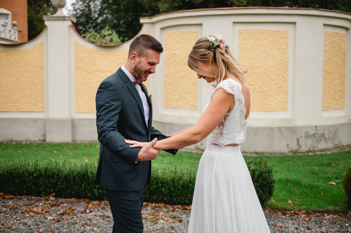 Ein Bräutigam im dunklen Anzug und eine Braut im weißen Kleid halten im Freien Händchen und lächeln sich an.