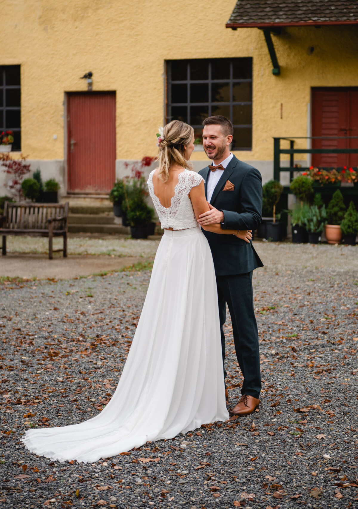 Braut und Bräutigam stehen draußen auf Kies, stehen einander gegenüber und lächeln. Die Braut trägt ein weißes Kleid und der Bräutigam einen dunklen Anzug. Im Hintergrund ist ein gelbes Gebäude mit einer roten Tür zu sehen.