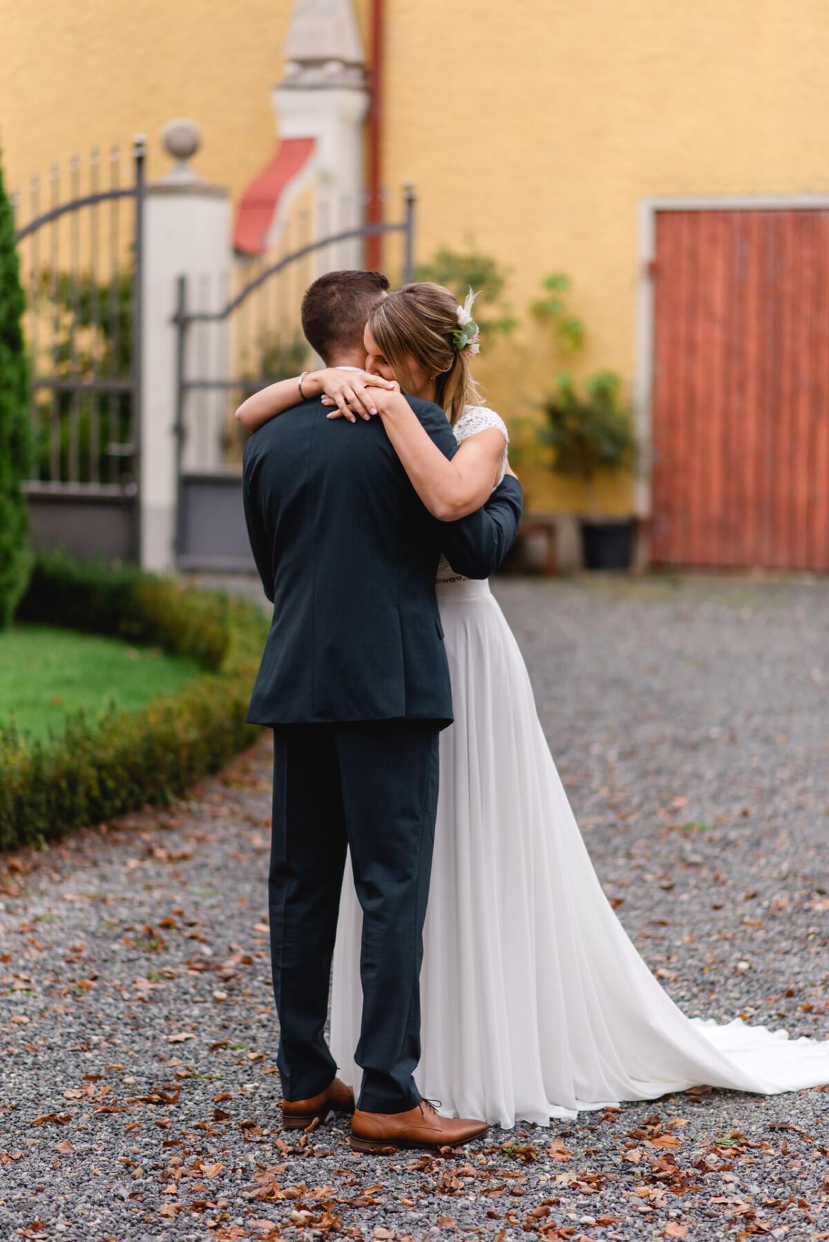 Ein Paar umarmt sich in einer Gartenkulisse. Die Braut trägt ein weißes Kleid und der Bräutigam einen dunklen Anzug. Sie stehen auf einem Kiesweg mit einem gelben Gebäude und einer Holztür im Hintergrund.