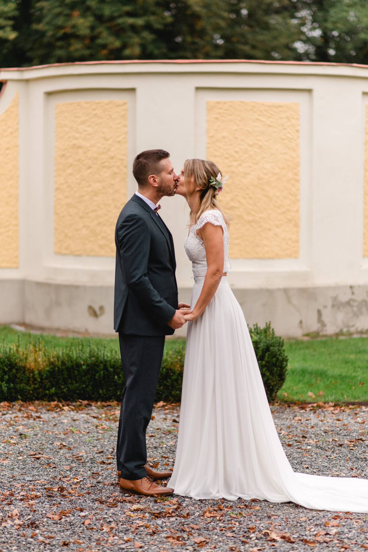 Ein Paar im Hochzeitskleid steht im Freien, hält Händchen und küsst sich, im Hintergrund eine gelb-beige Wand und Grünpflanzen.