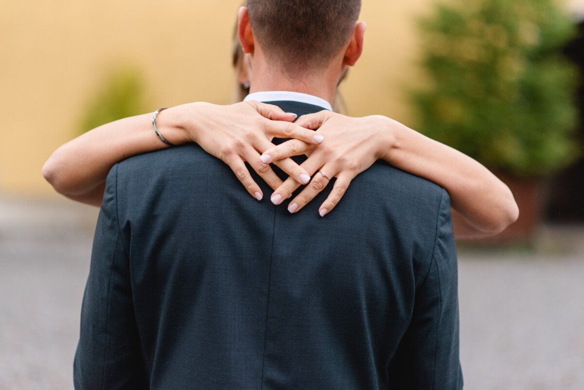Eine Person umarmt eine andere von hinten und hat die Hände um den Hals gelegt. Der Fokus liegt auf den Händen und der Rückseite des Anzugs der Person. Unscharfer Hintergrund mit Grün.