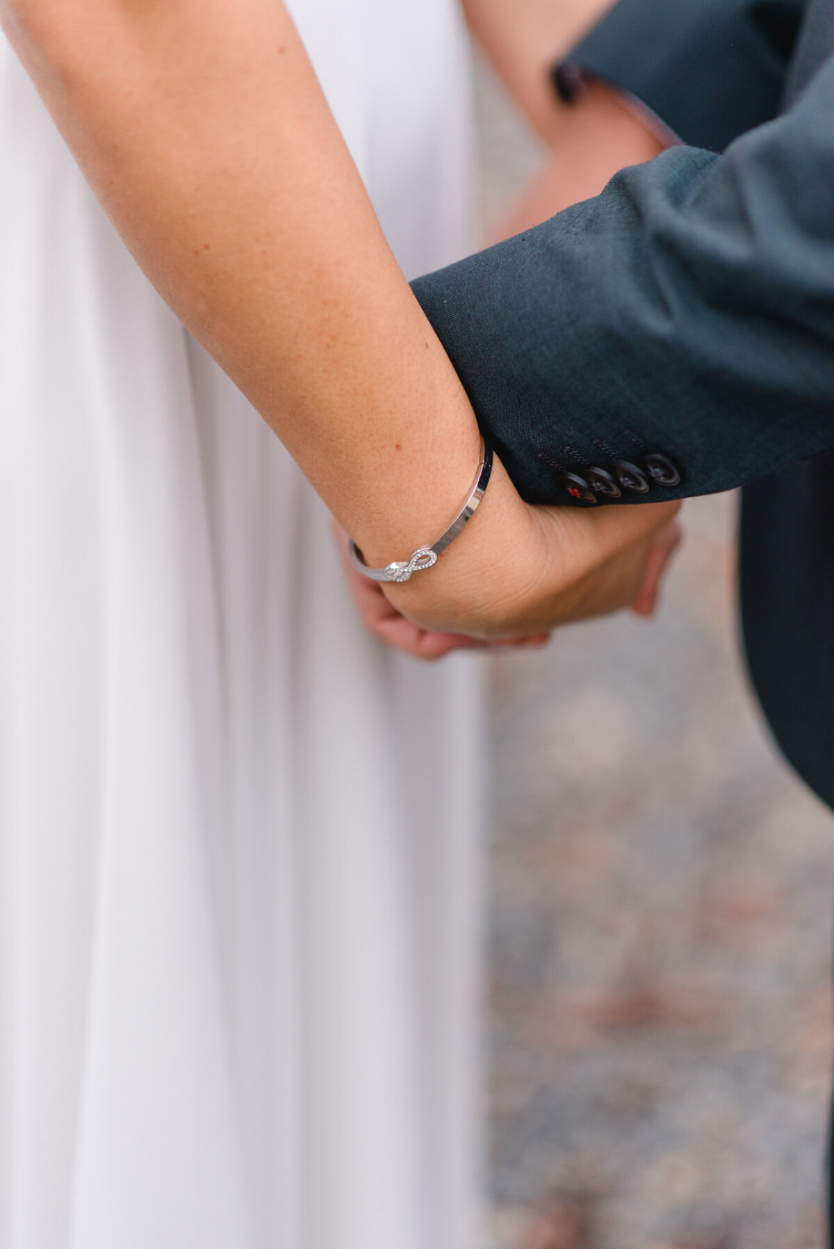 Nahaufnahme eines Paares, das Händchen hält; die eine trägt ein silbernes Armband und ein weißes Kleid, die andere ein dunkles Jackett.