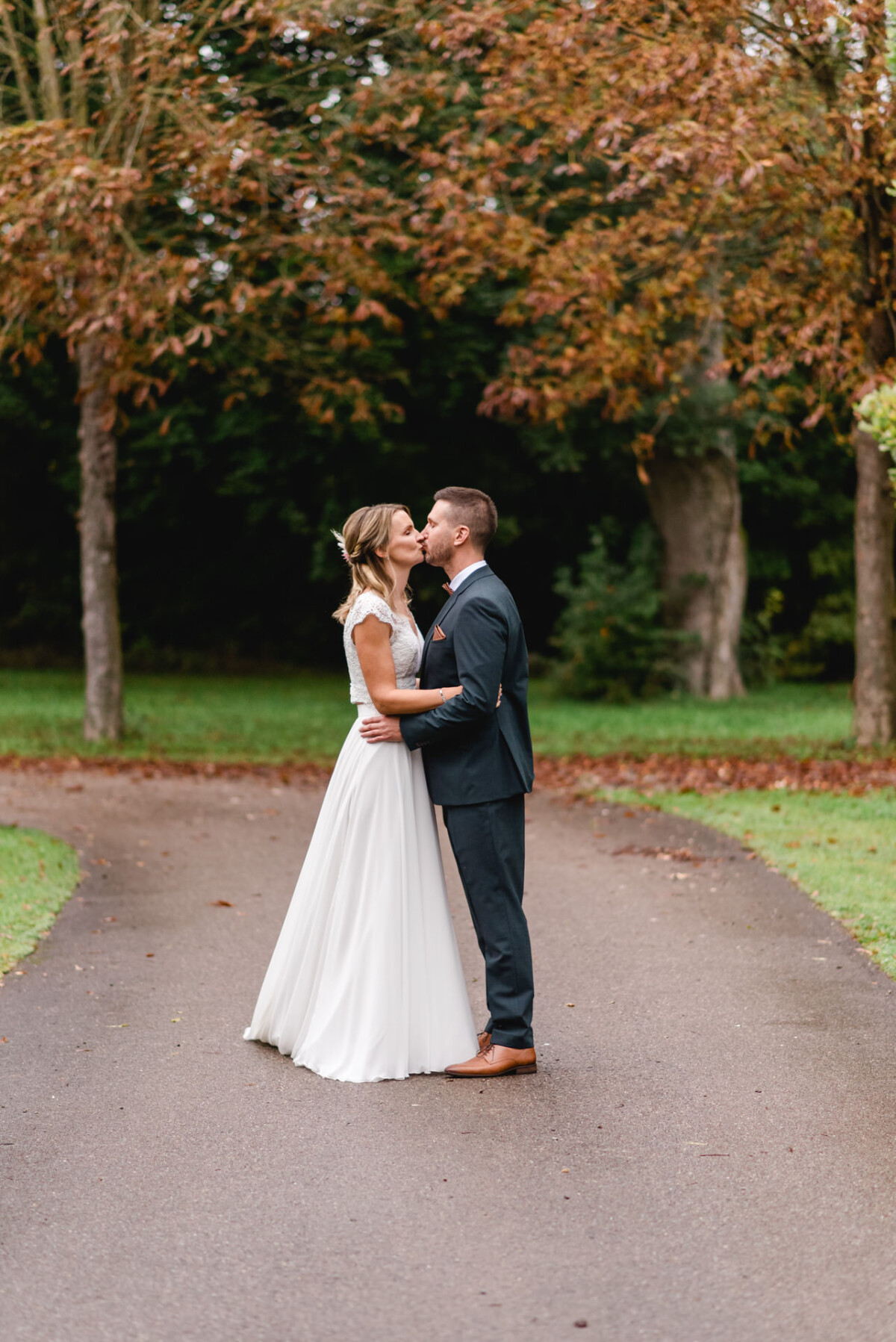 Ein Paar im Hochzeitskleid küsst sich auf einem von Bäumen gesäumten Weg in einem Park, umgeben von Herbstlaub.