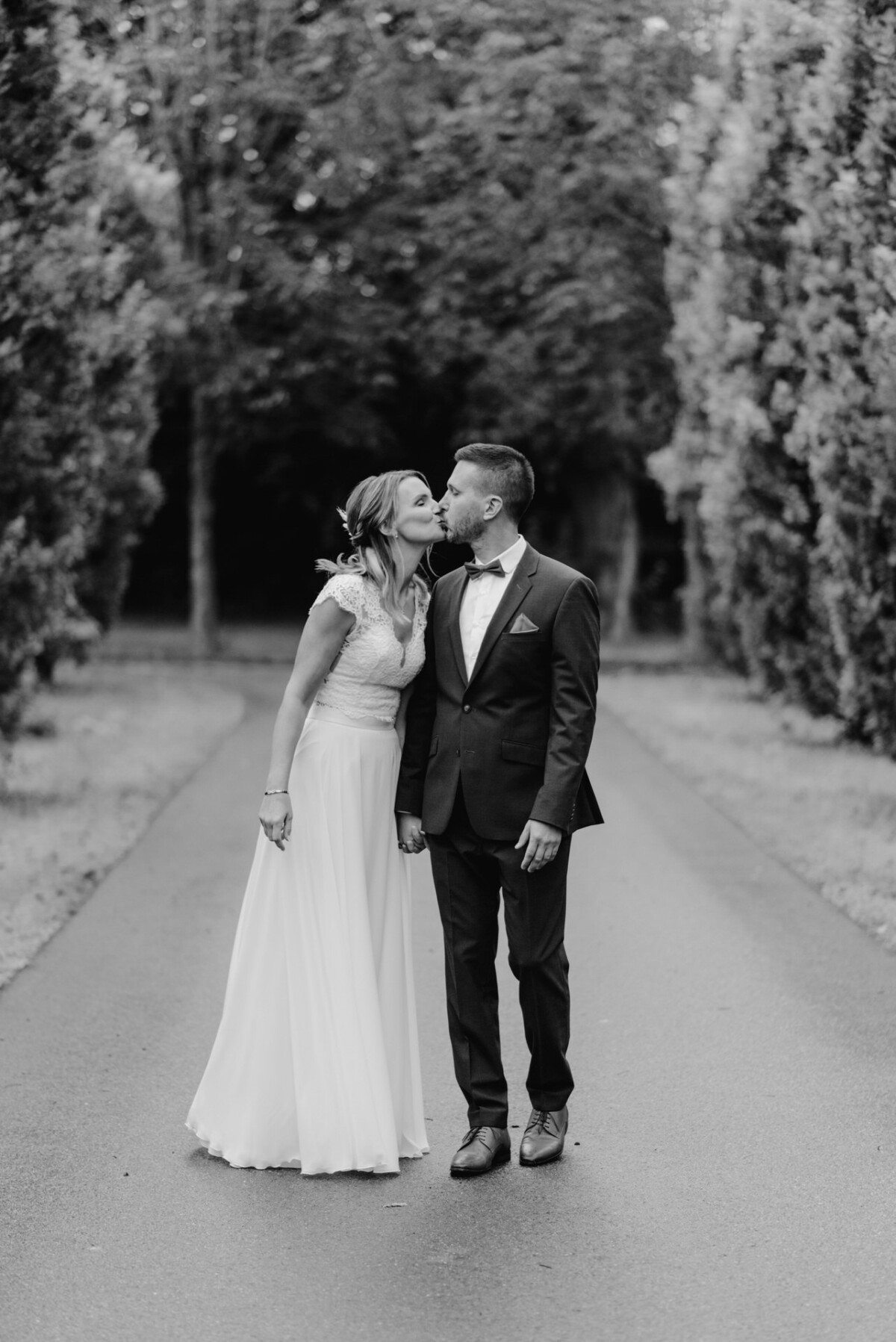 Braut und Bräutigam küssen sich, während sie auf einer von Bäumen gesäumten Straße stehen. Die Braut trägt ein weißes Kleid und der Bräutigam einen Anzug und eine Fliege. Das Bild ist schwarzweiß.