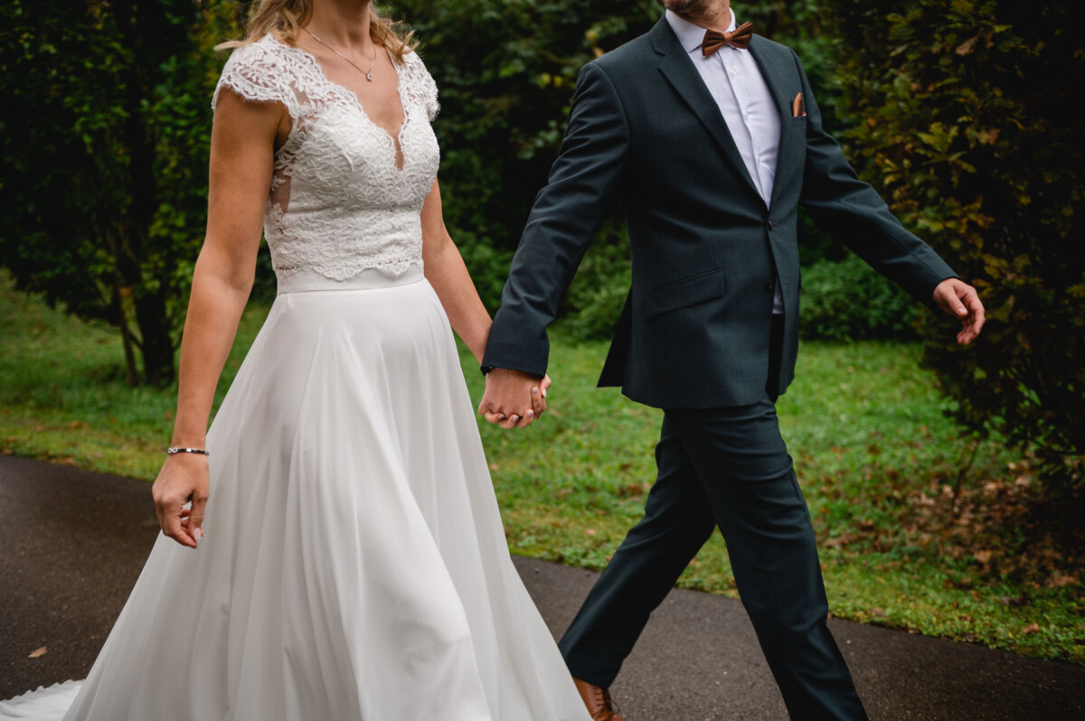 Eine Braut im weißen Kleid und ein Bräutigam im dunklen Anzug gehen Händchen haltend einen Weg entlang, umgeben von Grün.