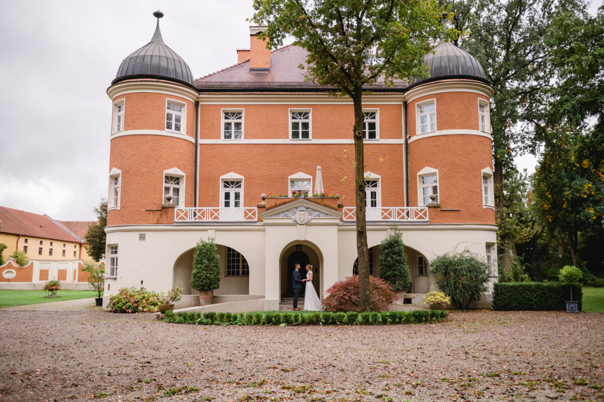 Braut und Bräutigam stehen vor einem historischen, rot-orangen Gebäude mit weißen Verzierungen und zwei Türmchen, umgeben von Grünflächen und einem Kieshof.