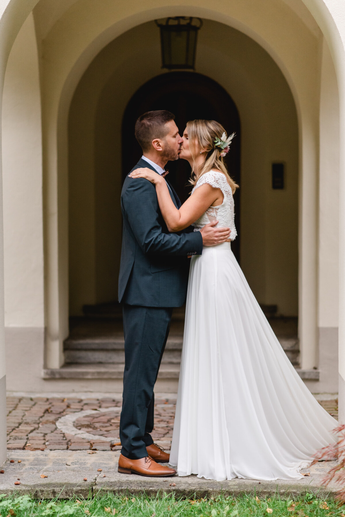 Braut und Bräutigam küssen sich vor einem Torbogen. Sie trägt ein weißes Kleid und er einen dunklen Anzug.