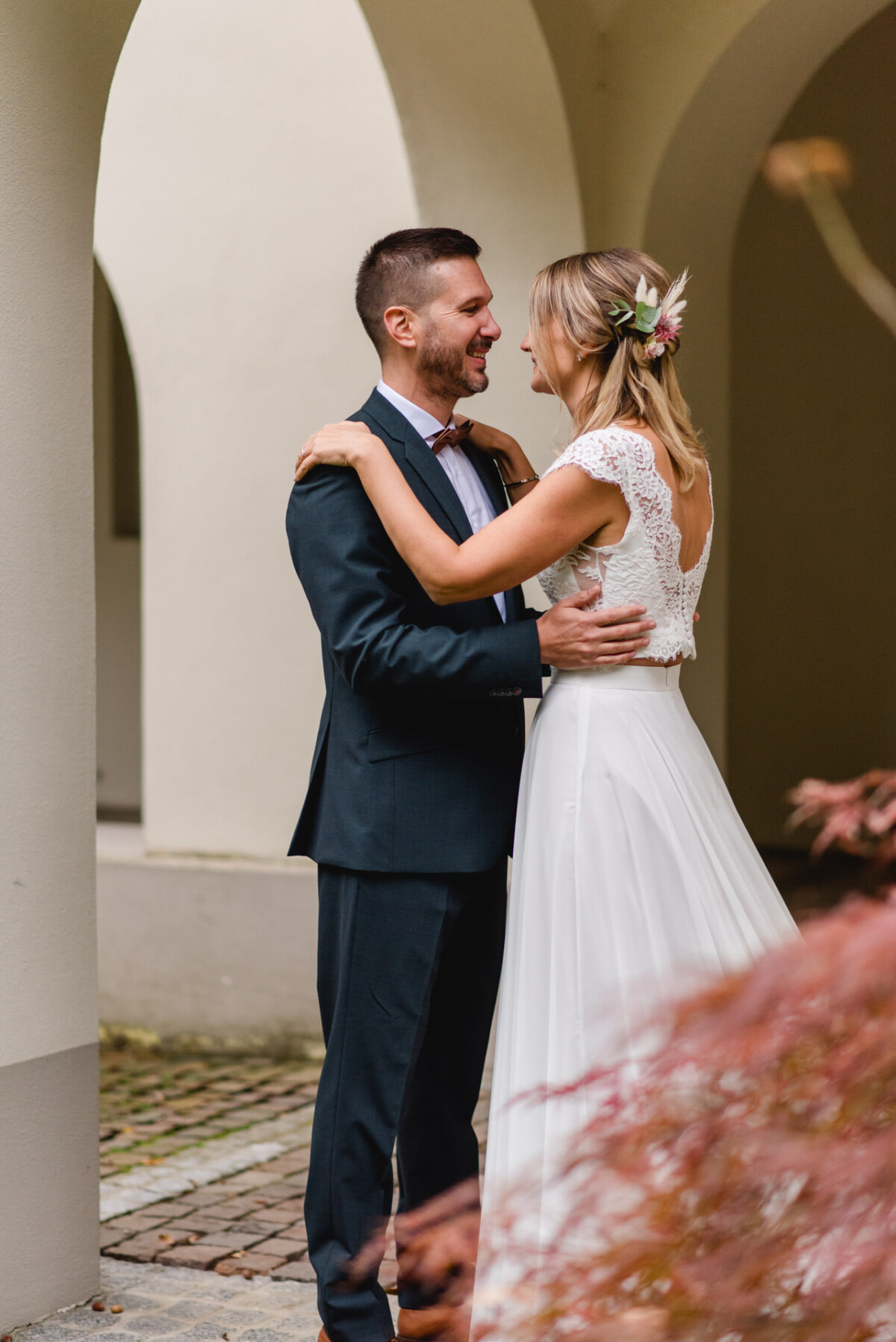 Ein Paar in formeller Hochzeitskleidung steht lächelnd und einander zugewandt unter einem Torbogen.