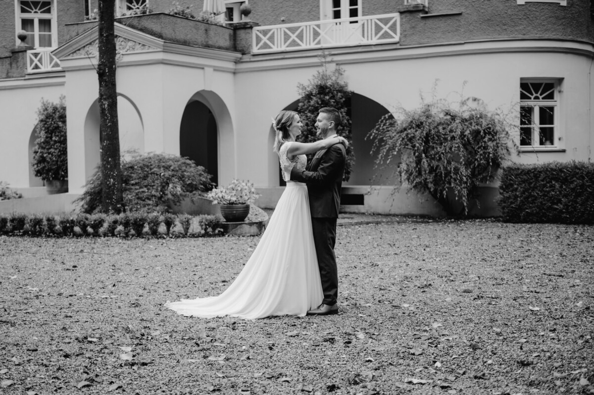Braut und Bräutigam umarmen sich im Freien vor einem großen Gebäude mit gewölbten Türen und üppigem Grün. Die Braut trägt ein langes, fließendes Kleid.