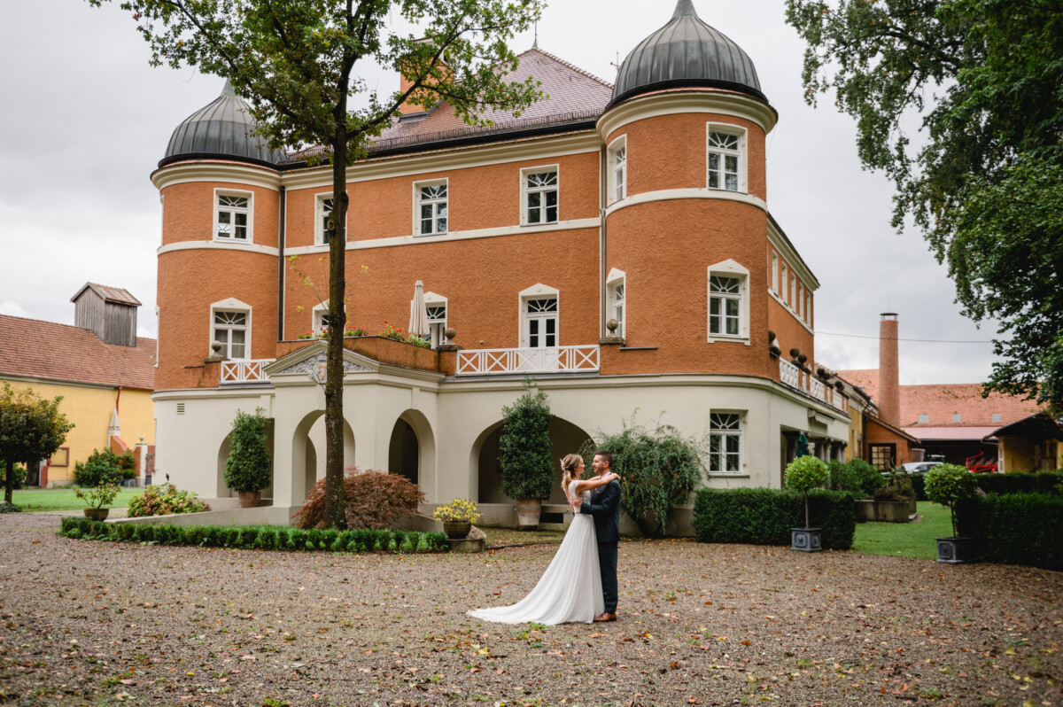 Ein Paar tanzt vor einem historischen orangefarbenen Gebäude mit weißen Akzenten und einem Baum in der Mitte, umgeben von einem Kieshof und Grünflächen.