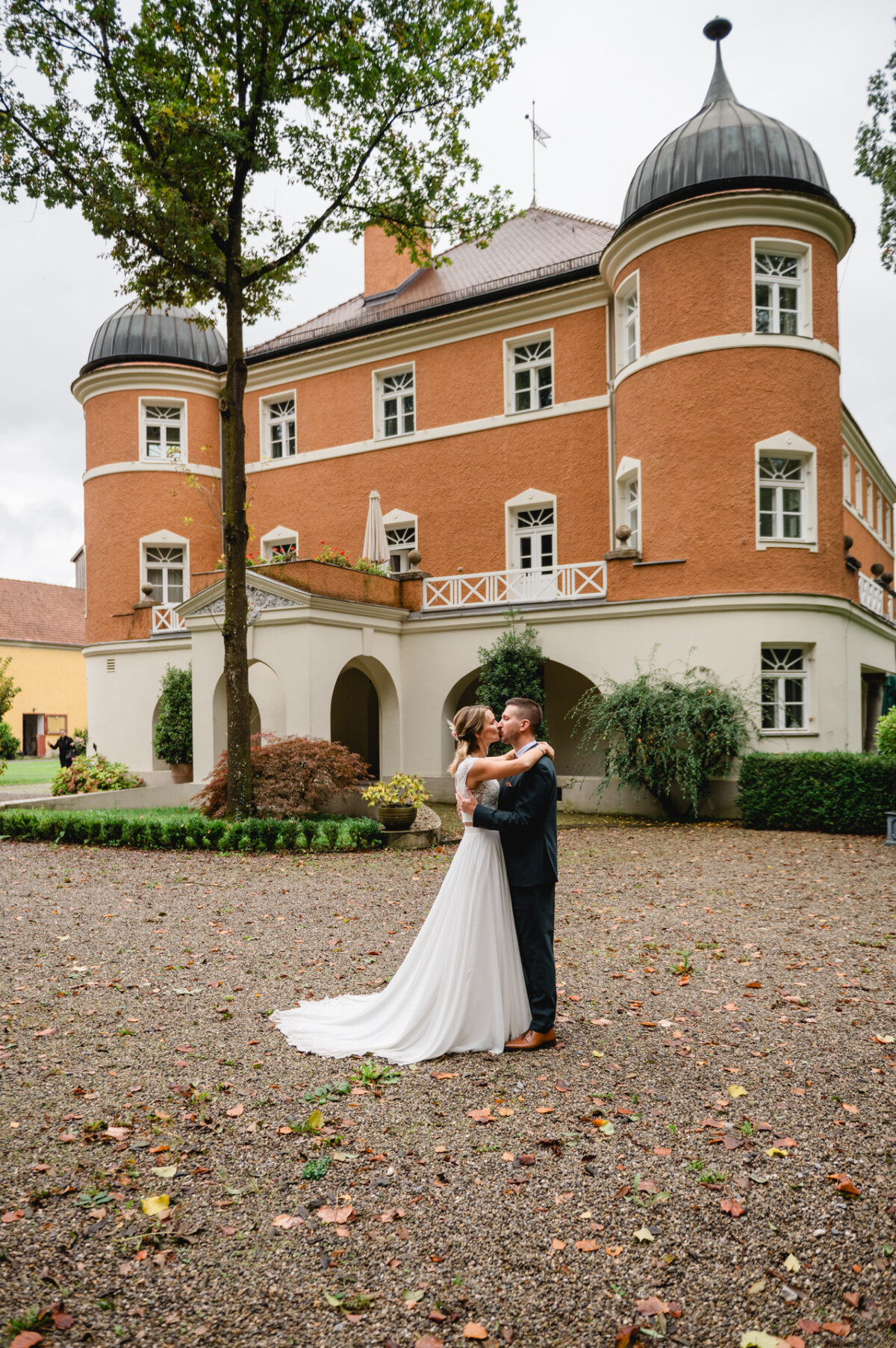 Ein Paar im Hochzeitskleid umarmt sich vor einem orangefarbenen Backsteingebäude mit Türmen und Bogenfenstern, umgeben von Bäumen und einem Kieshof.