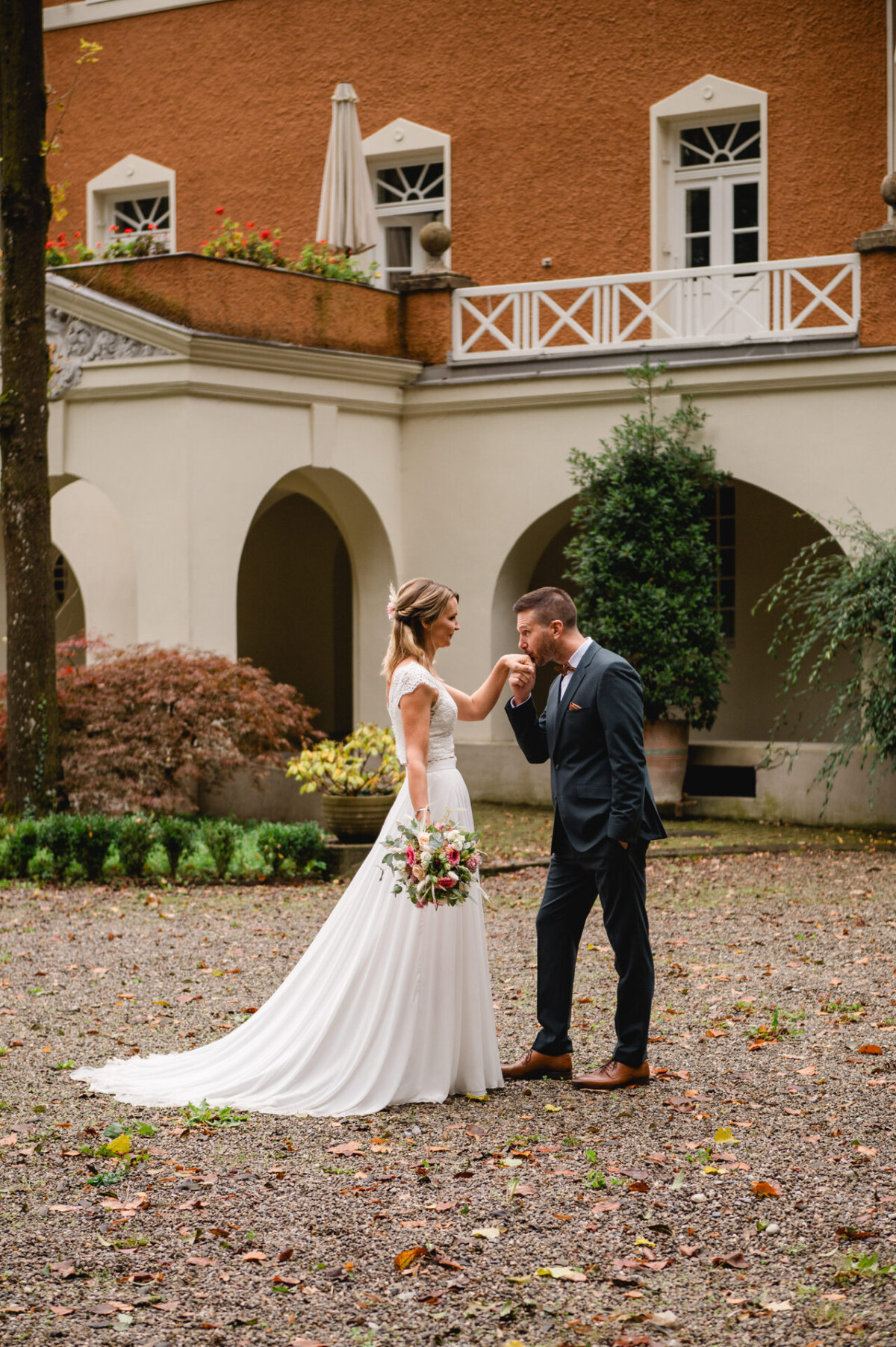 Braut und Bräutigam stehen in einem Innenhof; der Bräutigam küsst die Hand der Braut, während sie einen Blumenstrauß hält.