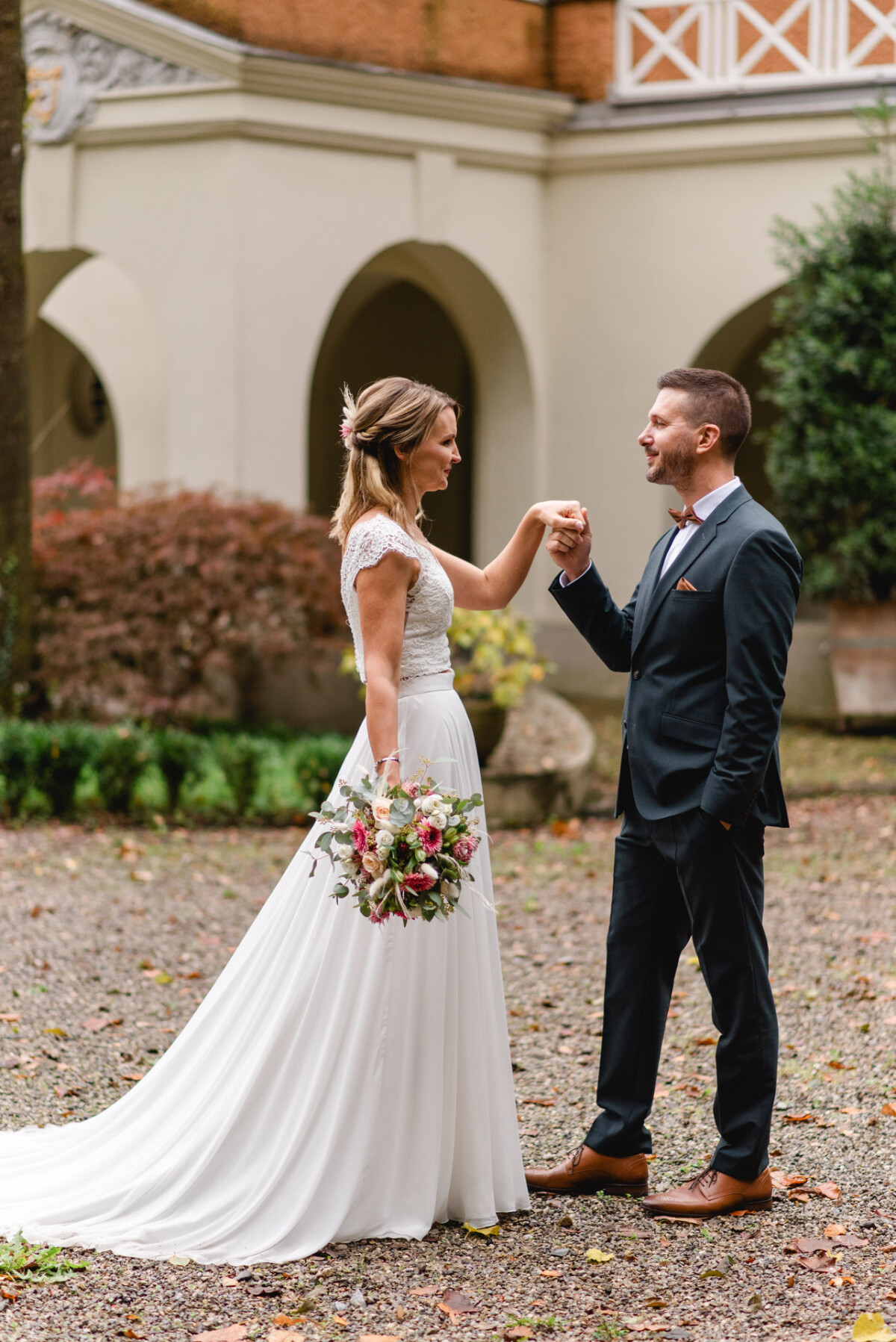 Ein Paar steht im Freien unter einem Torbogen. Die Frau, die ein weißes Brautkleid trägt und einen Blumenstrauß hält, und der Mann im schwarzen Anzug blicken einander an und halten Händchen.