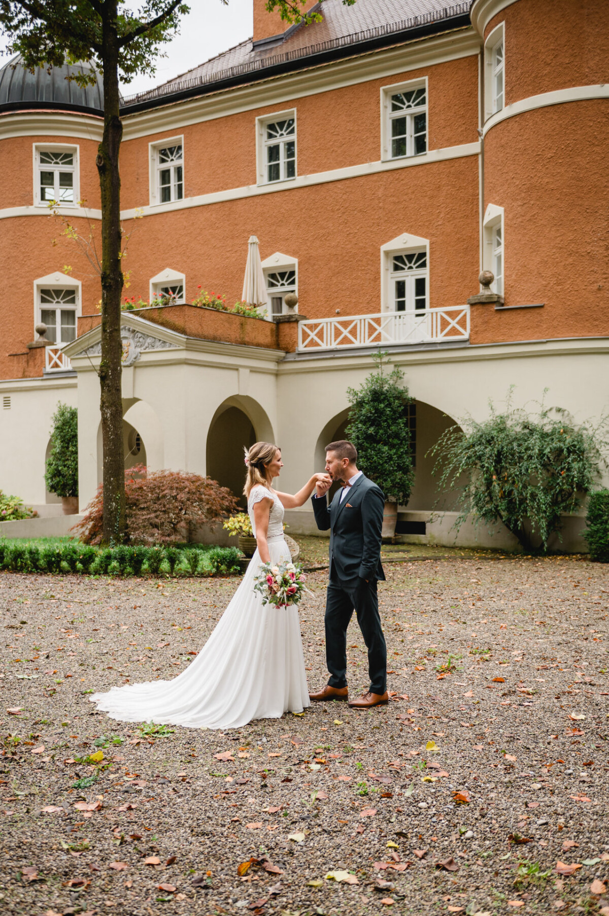 Braut und Bräutigam stehen auf einem Kiesweg vor einem großen Gebäude mit Bogenfenstern, umgeben von Grün. Der Bräutigam küsst die Hand der Braut.