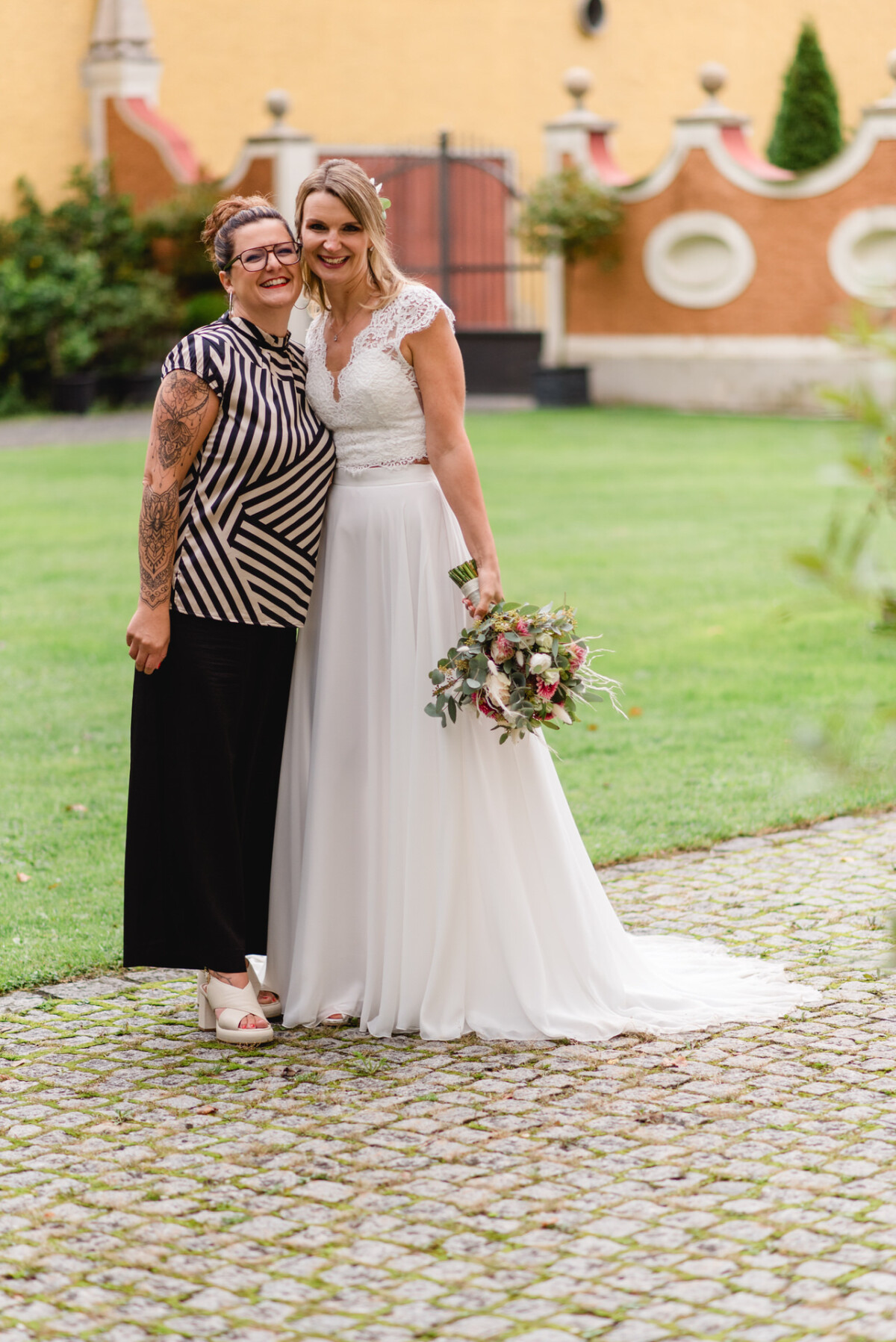 Zwei Frauen posieren im Freien auf einem Kopfsteinpflasterweg für ein Foto, eine in einem Brautkleid und mit einem Blumenstrauß in der Hand, die andere in einem schwarz-weiß gestreiften Oberteil.