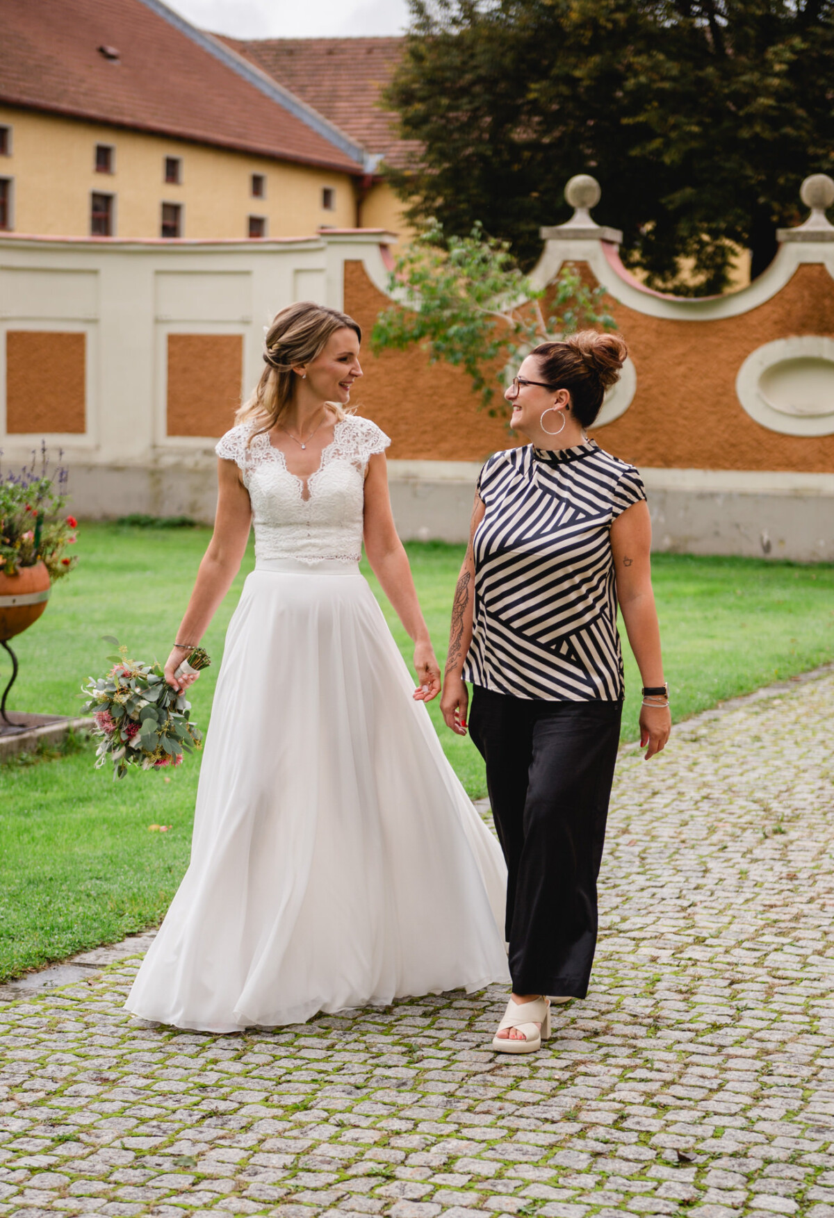 Eine Braut in einem weißen Kleid geht mit einer Frau in einem gestreiften Oberteil und schwarzen Hosen auf einem Steinweg in einem Garten spazieren.