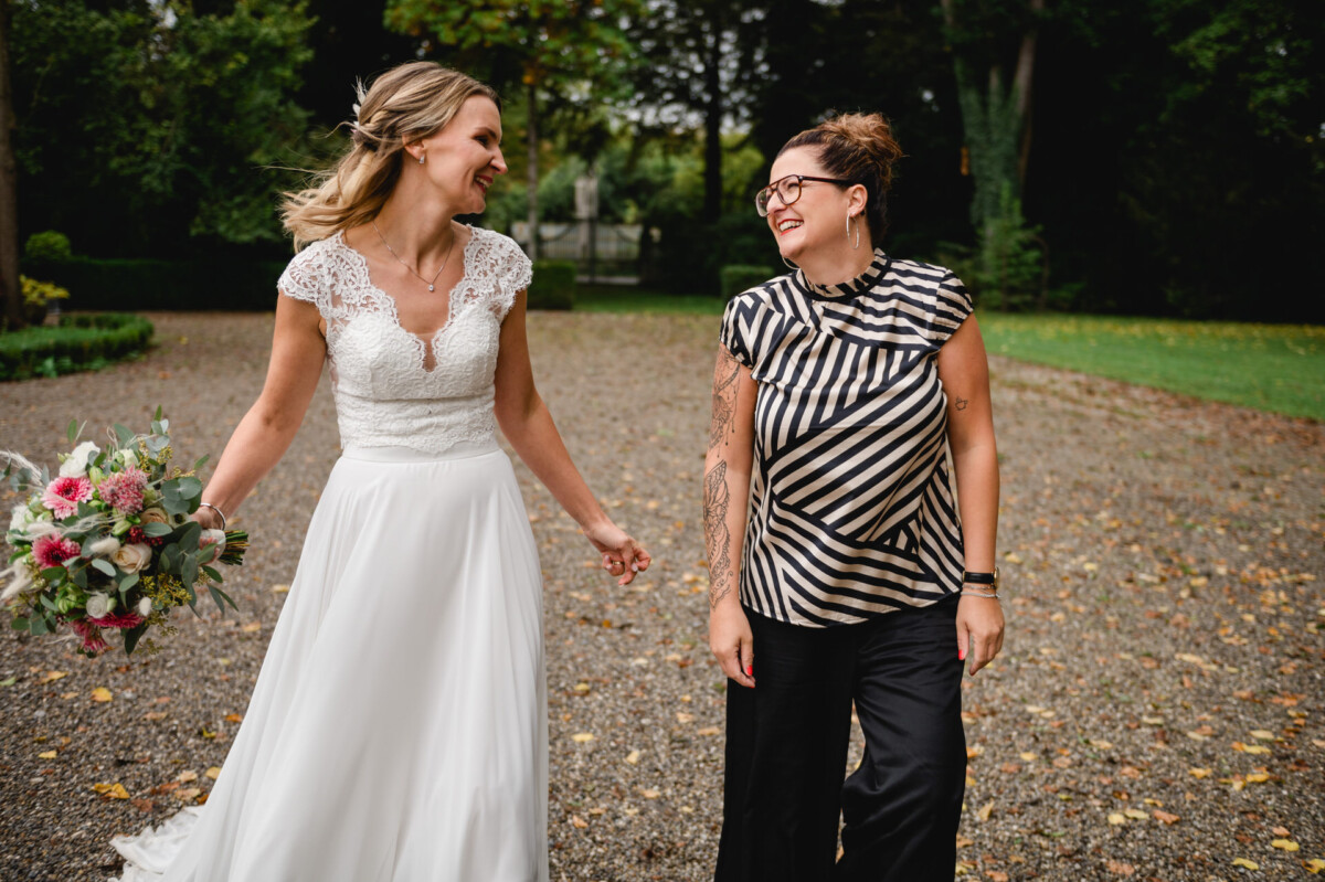 Eine Braut im weißen Kleid mit einem Blumenstrauß geht neben einer Frau im schwarz-weiß gestreiften Oberteil her. Beide lächeln auf einem von Grün umgebenen Kiesweg.